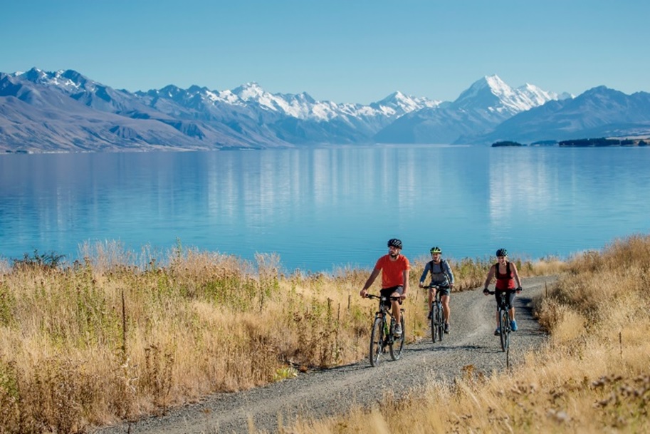 An image of three people on bicycles, cycling on a track with New Zealand mountains and a lake in the background.. 