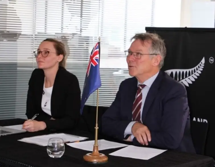 An image of David Parker and Alison Hamilton sitting at a desk with an NZ flag ornament prominently set on the table in front of them..