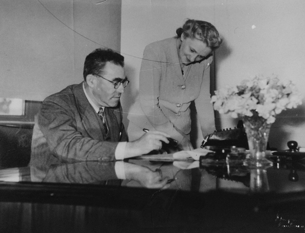 A historical image of Carl Berendsen, sitting at a desk holding a pen with papers in front of him. There is a woman standing to his left, looking at the papers on the desk.. 