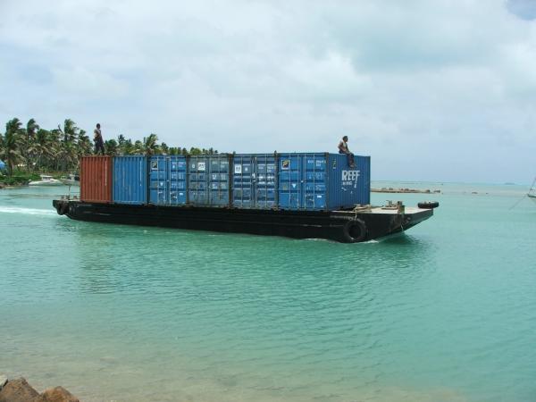 An image of a barge with six shipping containers on it, and a man sitting atop one of the shipping containers. . 