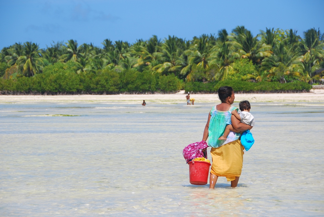 An image of a woman in the pacific islands, walking through shallow sea water with a young child balanced on her hip and a bucket in her other hand. . 