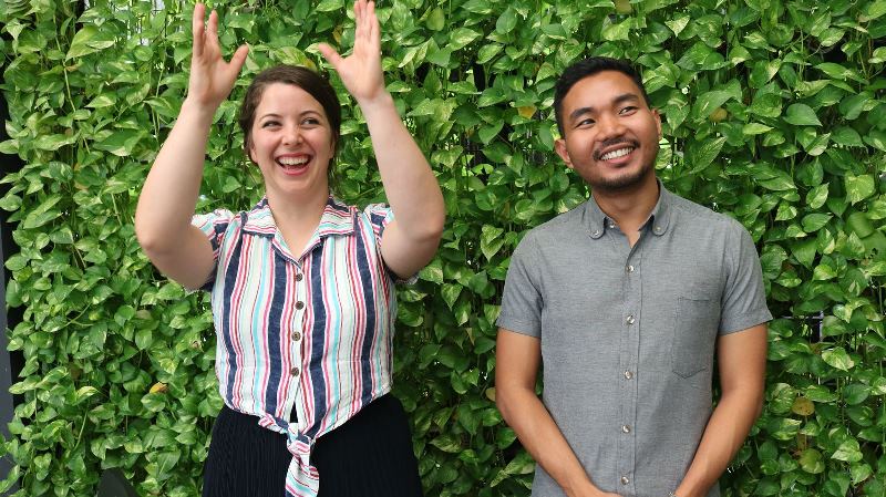 An image of two people, a man and a woman, standing in front of a lush hedge, posing for a photo with smiles on their faces.. 